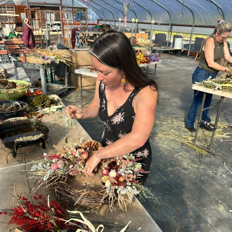 Woman arranging flowers in a greenhouse setting with tables and plants in the background.