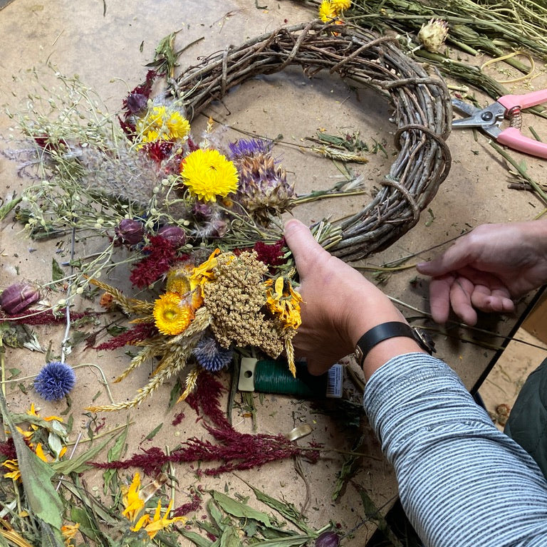 Person crafting a wreath with dried flowers and leaves on a wooden surface.