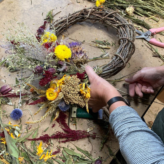 Person crafting a wreath with dried flowers and leaves on a wooden surface.