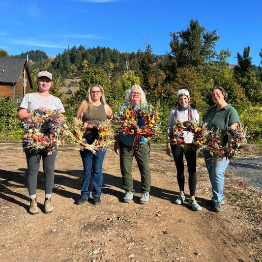 Five women holding dried flower bouquets outdoors with a clear blue sky and greenery in the background.
