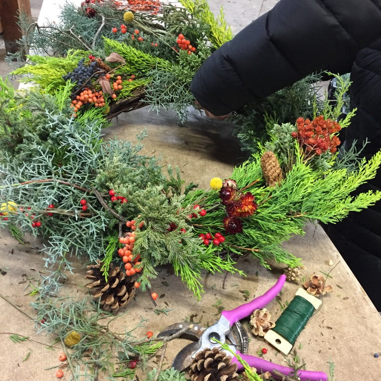 Person arranging a festive wreath with greenery, berries, and pinecones on a table.