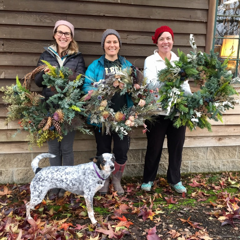 Three women holding wreaths with a dog in front of a wooden building.