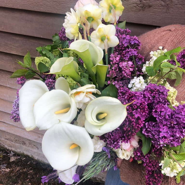 Bouquet of white calla lilies and purple flowers held by a person against a wooden wall.